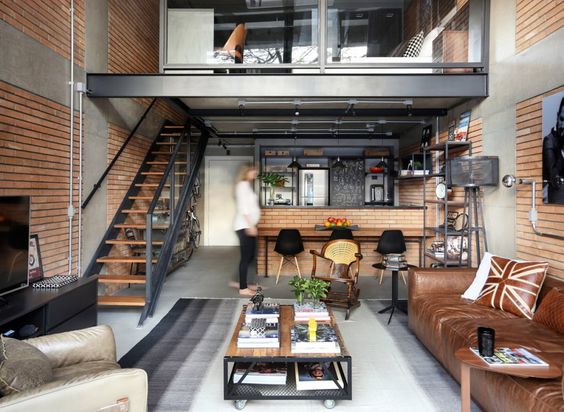 Loft com pé-direito duplo com decoração totalmente industrial, contando com parede de tijolos e sofá de couro marrom.