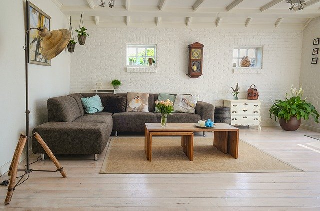 Sala de estar com sofá, almofadas, luminária e mesa. Está decorada com plantas, flores, quadro, relógio e tapete.