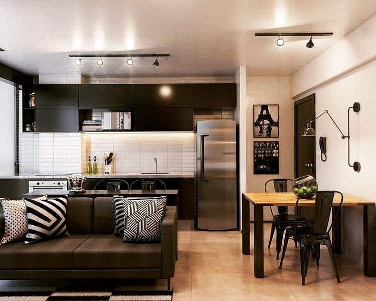Flat com cozinha integrada a sala de estar. Na imagem é possível ver a geladeira, a pia, os armários, o sofá e uma mesa de canto com cadeiras e iluminação.