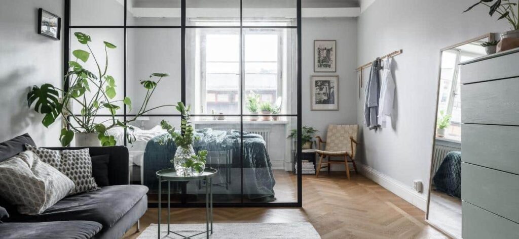 Kitnet com sala de estar e quarto. O espaço está otimizado por meio do uso de cores claras e separadores de vidro.