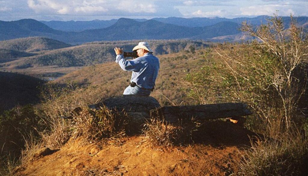 O fotógrafo Sebastião Salgado encontra-se sentado em cima de uma pedra, no meio de uma paisagem árida e montanhosa. Esta é uma cena do filme O Sal da Terra.