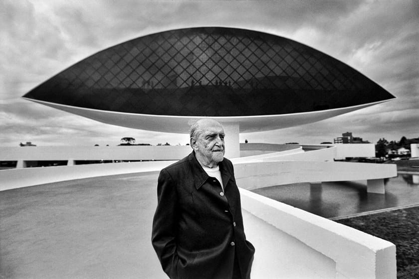 Fotografia de Oscar Niemeyer em preto e branco. Ao fundo o museu lembra o formato de um olho, suspenso em um caminho curvilíneo.