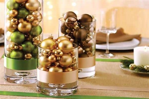 Decoração feita com bolas de Natal em vasos de vidro (ou copos largos), sobre uma mesa.