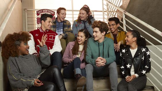 Atores da continuação de High School Musical sentados juntos em uma escada da famosa escola East High. A nova série estará disponível com exclusividade no Disney+.