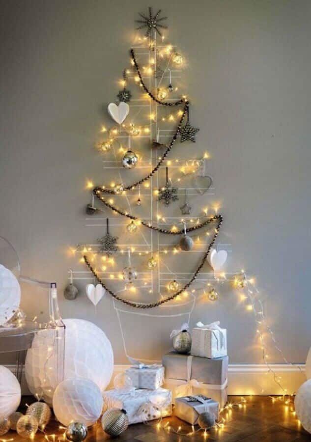 Árvore de Natal desenhada com giz numa parede cinza, e decorada com pisca-piscas e alguns outros enfeites como bolas de Natal, corações e penduricalhos.