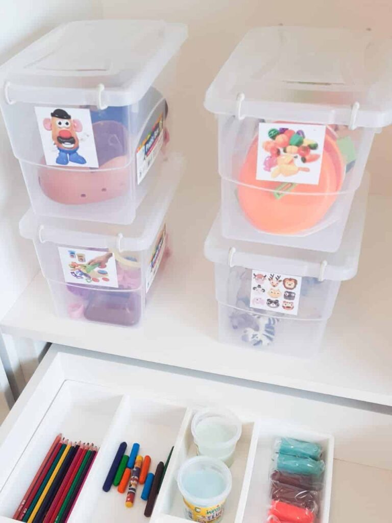 Caixas transparentes organizadoras armazenam brinquedos e materiais de arte como lápis colorido e massa de modelar colorida.