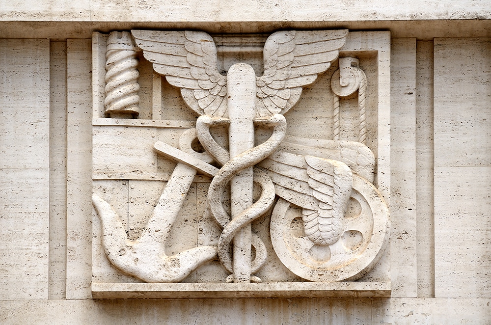 Fotografia do símbolo do comércio, localizado na fachada. O símbolo é um bastão em torno do qual se entrelaçam duas serpentes.