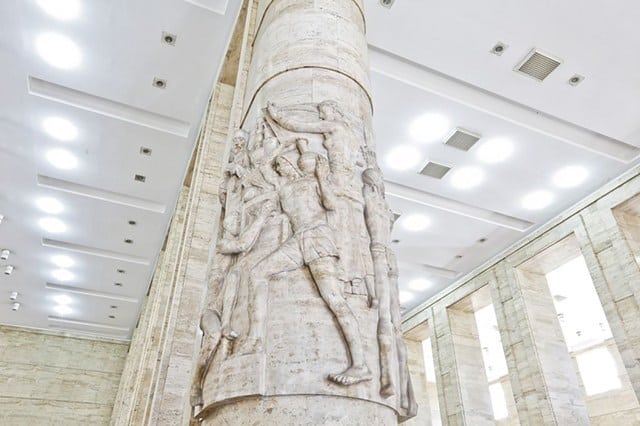 Fotografia de uma das colunas do interior do edifício, com escultura entalhada que representa trabalhadores carregando objetos.