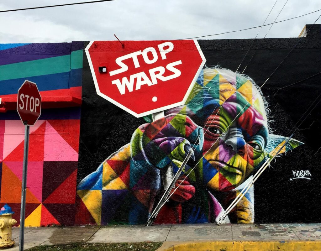 Em um fundo preto, com uma padronagem bem colorida, Mestre Yoda segura uma placa vermelha escrita: “Stop Wars”.