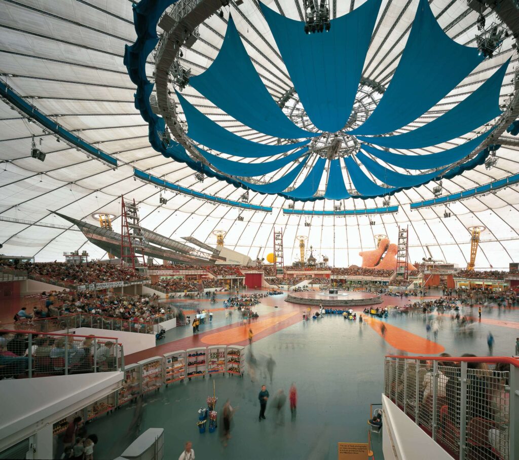 O interior do Millenium Dome é amplo e bastante utilizado para eventos. Nessa imagem, há muitas pessoas sentadas em arquibancadas e, ao centro, um palco em formato circular.