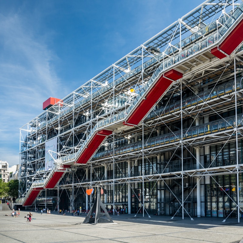 O vidro e o aço do Centro Pompidou refletem com a luz do sol, destacando o edifício da paisagem. A escada rolante fica na parte externa e possui pintura na cor vermelha.