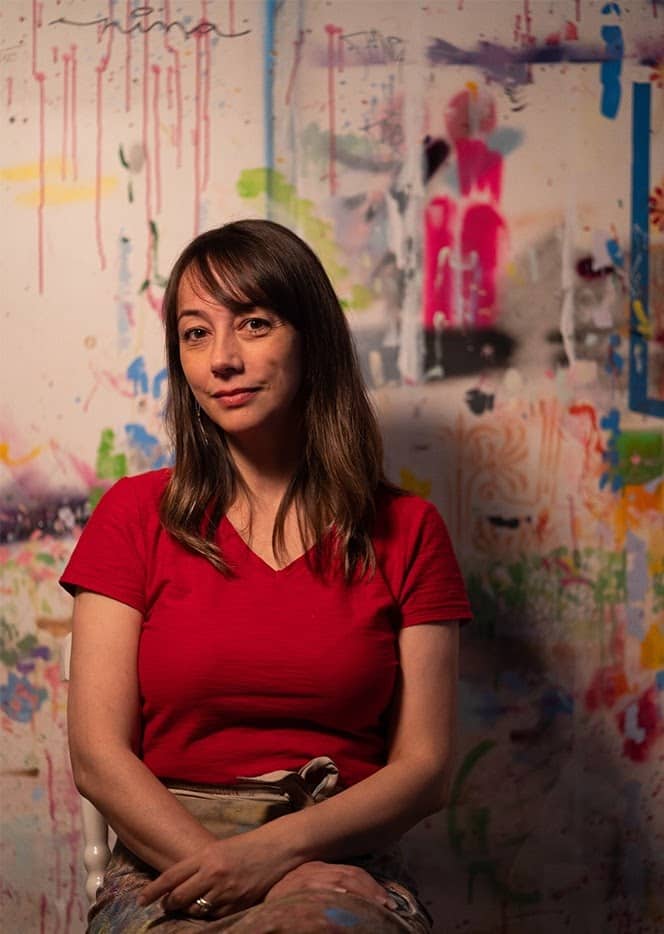 Nina Pandolfo posa em frente a uma parede com algumas intervenções artísticas coloridas e seu nome assinado.