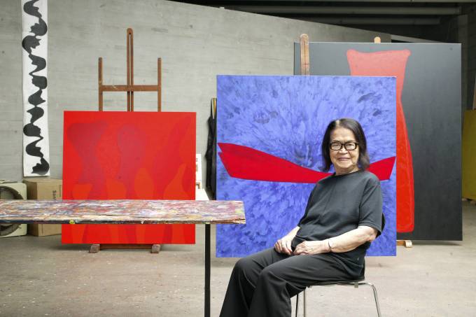 Artista Tomie Ohtake sentada em primeiro plano. Ao fundo, suas obras compõem um cenário em tons de vermelho e azul.