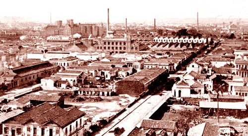 Imagem antiga do Brás mostrando casas baixas com telhado de barro e diversas fábricas com chaminés.