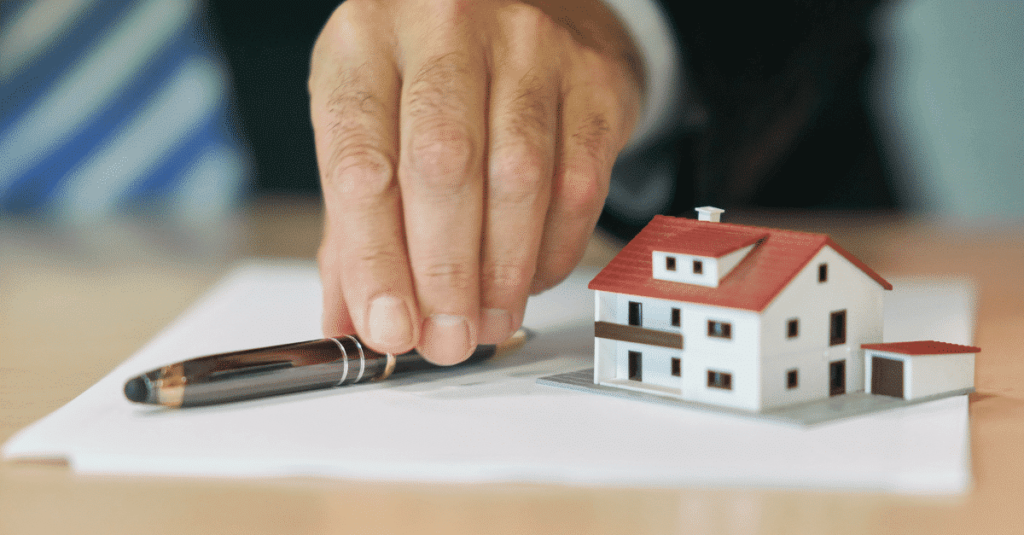 Mão segurando uma caneta sobr eum papel, ao lado de uma casinha, representando o ato de investir em imóveis.
