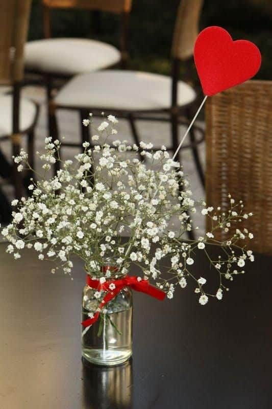 Arranjo de flores brancas em um pequeno vaso de vidro, decorado com laÃ§o vermelho e coraÃ§Ã£o.
