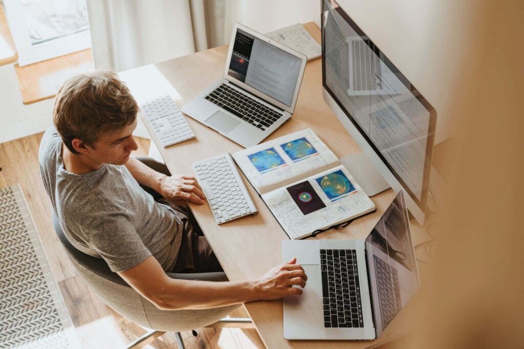 Homem trabalhando em seu escritório em casa: mesa com um computador e dois notebooks, acompanhado de dois teclados e um livro.