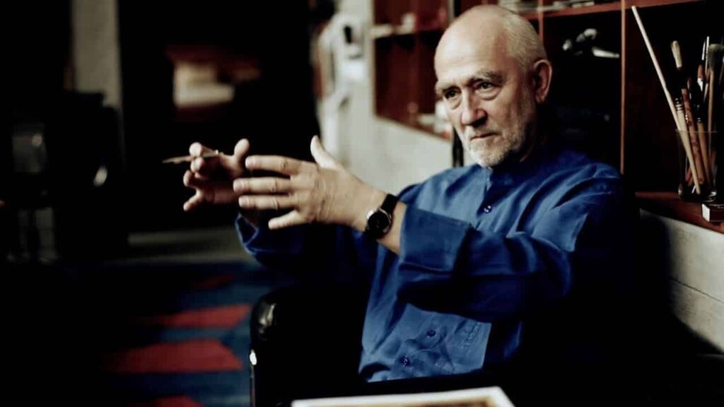 Peter Zumthor gesticula, enquanto está sentado em uma poltrona preta, veste uma camisa azul escuro, com um relógio no pulso esquerdo e segura uma caneta na sua mão direita.