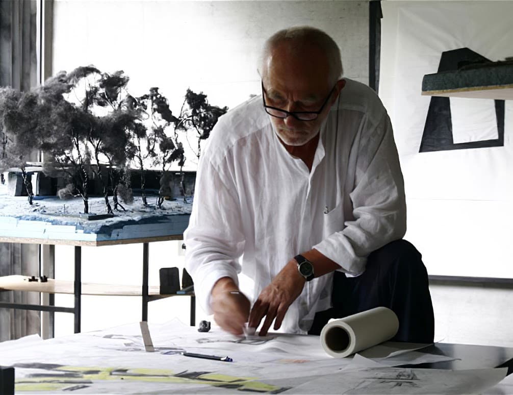 Peter Zumthor, de camisa branca, relógio no pulso esquerdo e óculos, posicionado em frente a uma mesa com papéis e canetas, trabalha em mais um de seus projetos.
