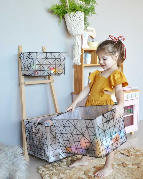 Criança com um vestido cor mostarda segurando uma caixa transparente com detalhes em preto guardando os brinquedos.