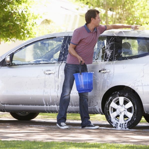Homem usando uma blusa listrada roxa e branco, calça jeans e tênis, lava um carro prata de porte médio, utilizando um balde com água e sabão.