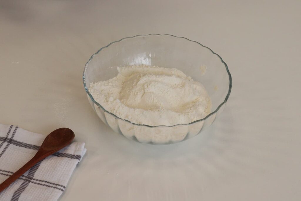 Ingredientes secos dentro do bowl, ao lado de uma colher de pau em cima de um pano de prato quadriculado sobre a mesa.