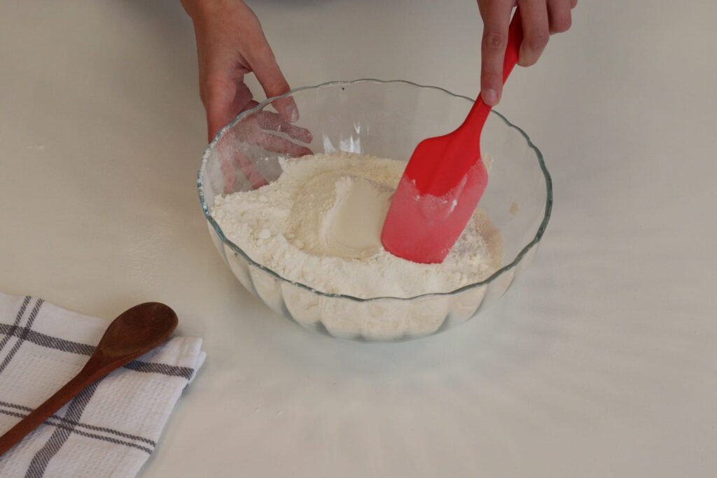 Ingredientes secos do bolo de cenoura dentro do bowl, sendo misturados com uma espátula vermelha, ao lado de uma colher de pau em cima de um pano de prato quadriculado sobre a mesa.