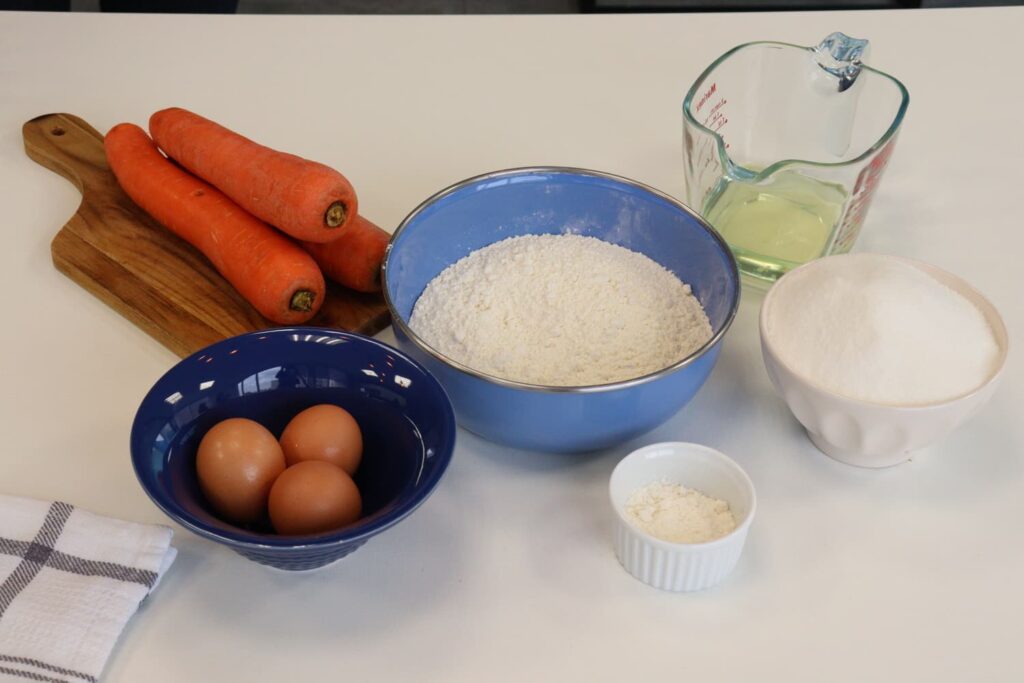 Ingredientes necessários para a massa do bolo de cenoura sobre a mesa: três cenouras, três ovos, açúcar, fermento químico, óleo de canola e farinha de trigo.