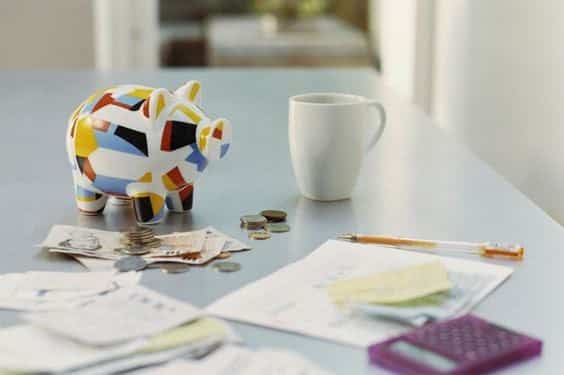 Cofre de porquinho colorido, moedas, contas, caneta, calculadora e notas fiscais, ao lado de uma caneca sobre a mesa.